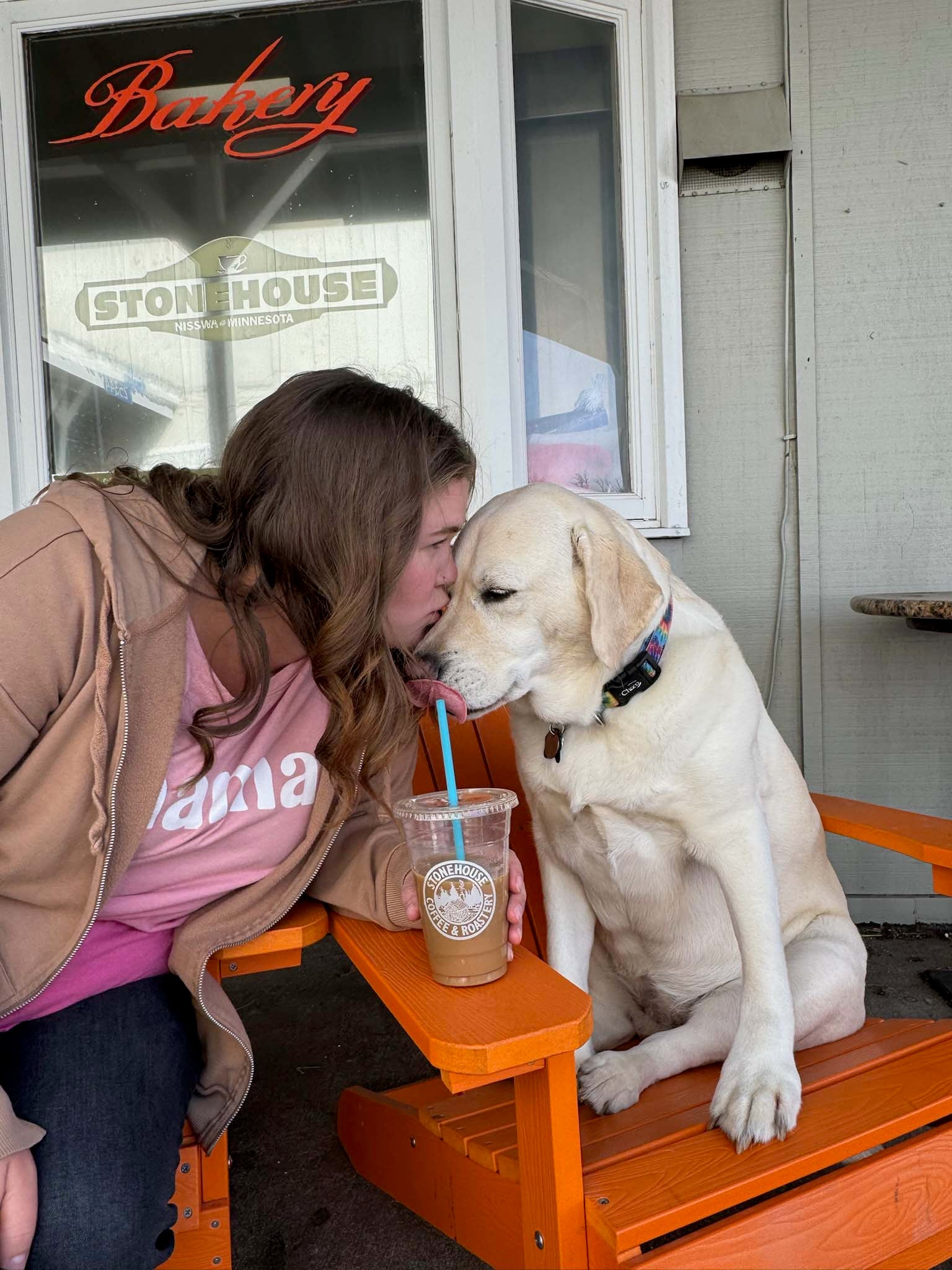 Woman and dog sitting on an orange bench in front of a bakery.